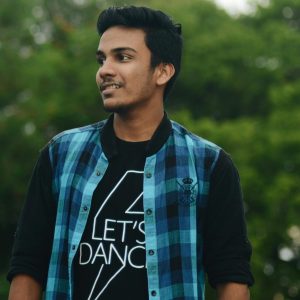 A young man in a casual plaid shirt smiling while standing outside against greenery.